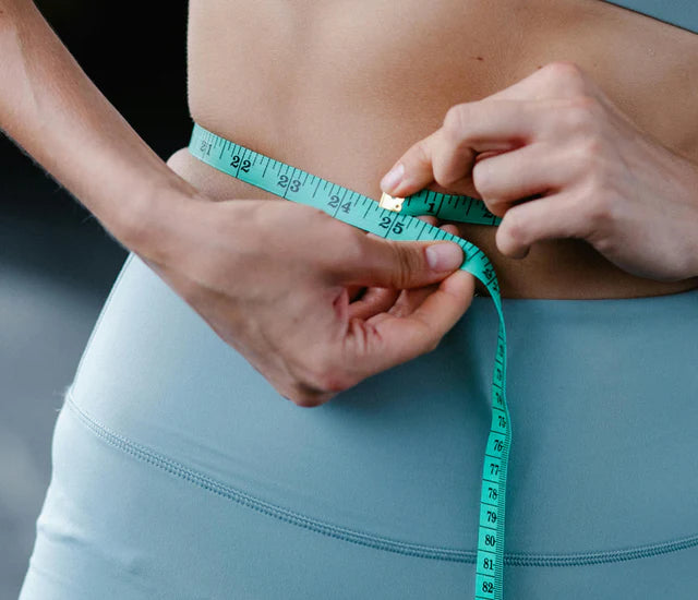 A person wearing blue exercise leggings and a matching sports bra uses a teal measuring tape to measure their waist.