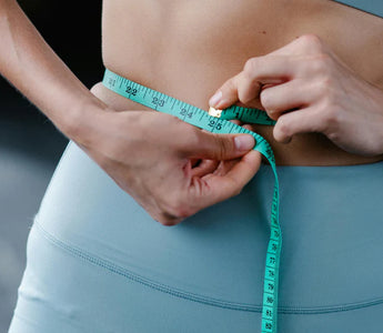 A person wearing blue exercise leggings and a matching sports bra uses a teal measuring tape to measure their waist.