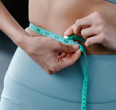 A person wearing blue exercise leggings and a matching sports bra uses a teal measuring tape to measure their waist.